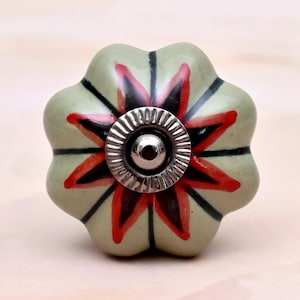 May include: A flower-shaped ceramic cabinet knob with a sage green base colour. The surface features a painted star pattern in red and black radiating from the centre. A circular, ridged silver-toned metal hardware piece is fixed in the middle.