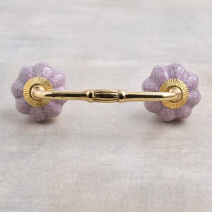 May include: A pair of ceramic drawer pulls in a light purple color with a textured finish. The pulls have a gold metal base and a curved shape.