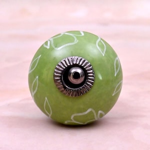 Op de afbeelding: Een ronde, lichtgroene keramische meubelknop met een handgeschilderd wit bladpatroon. Het midden van de knop is voorzien van een cirkelvormig, zilverkleurig metalen beslag met een geribbelde rand en een centraal gat voor montage.