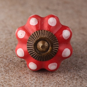 May include: A red ceramic cabinet knob with white polka dots. The knob has a flower-like shape with a brass-colored center. This decorative hardware is suitable for drawers and cabinets.