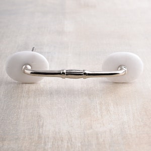 Op de afbeelding: Witte keramische ladegreep met een zilveren metalen staaf. De greep heeft een eenvoudig, elegant ontwerp.