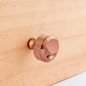 May include: A round, copper-colored cabinet knob with a ridged design. The knob is mounted on a light wood surface.