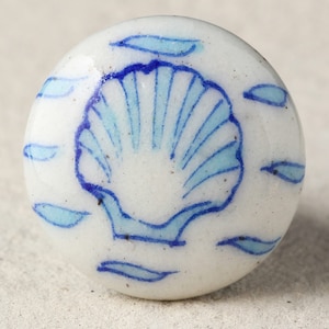 Op de afbeelding: Witte keramische kastknop met een blauw zeeschelpontwerp. De zeeschelp staat centraal, omringd door blauwe, bladachtige accenten. Deze decoratieve knop voegt een maritieme sfeer toe aan meubels.