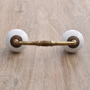 May include: White ceramic drawer pulls with a brass bar connecting them. The pulls have a decorative design around the edge.