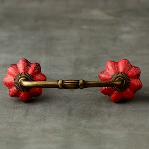 Pode incluir: Um puxador de gaveta com dois botões de cerâmica vermelhos em forma de flor com manchas pretas, ligados por uma pega central de metal em tom de latão antigo. As ferragens incluem placas decorativas em tom de latão na base.