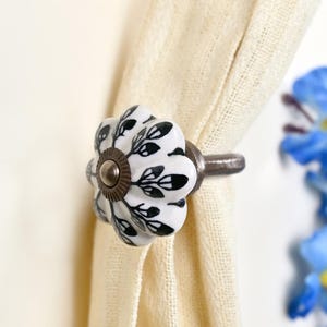 May include: A white ceramic curtain tieback featuring a scalloped, flower-like shape with a black leaf pattern. The center has a circular bronze-toned metal accent, and a matching bronze-toned metal post extends from the back to secure the fabric.