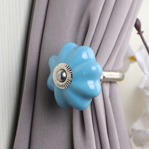 May include: A light blue ceramic curtain tieback with a silver metal ring. The tieback is shaped like a flower with a raised center.