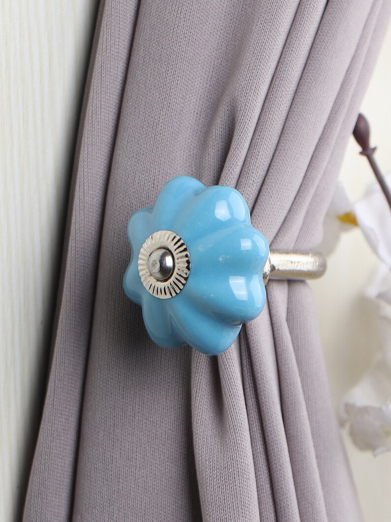 May include: A light blue ceramic curtain tieback with a silver metal ring. The tieback is shaped like a flower with a raised center.