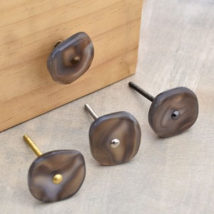 Pode incluir: Quatro botões de armário com um design de ágata cinza e detalhes metálicos. Os botões têm uma forma quadrada com bordas arredondadas. Os botões são mostrados em uma superfície de madeira e uma superfície cinza.