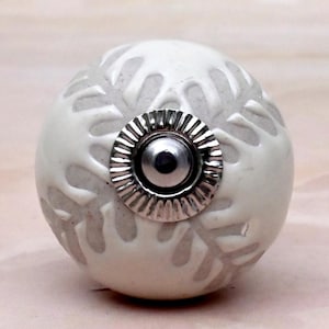 May include: A spherical cabinet knob featuring a white, raised, leaf-like pattern over a light gray, textured background. The center of the knob is adorned with a circular, metallic silver-toned hardware piece with a fluted, ridged edge.