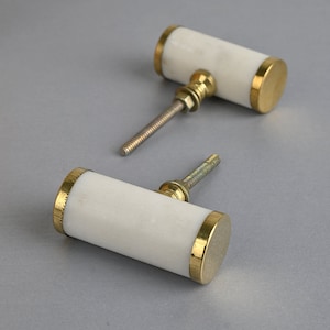 Könnte beinhalten: Zwei weiße Marmorknaufgriffe mit goldenen Metallakzenten. Die Knöpfe sind zylindrisch geformt und haben eine Schraube auf der Rückseite.