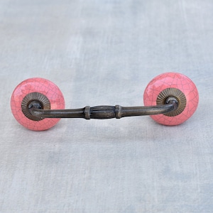 Pode incluir: Um puxador de gaveta com dois botões redondos de cerâmica rosa com acabamento em esmalte craquelado escuro. Os botões são ligados por uma alça de metal curva em tom de bronze antigo com um detalhe central canelado e bases metálicas estriadas.