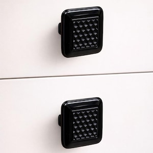 May include: Two black, square drawer pulls with a diamond pattern are mounted on a white drawer. The drawer has a wooden top. Each pull is approximately 4 cm square.