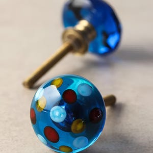 Op de afbeelding: Twee blauwe glazen kastknoppen. Eén knop is effen blauw, de andere heeft een stippenpatroon met rode, gele en witte stippen.