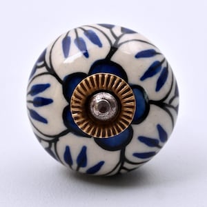 May include: A round ceramic cabinet knob featuring a hand-painted floral pattern in deep blue and black on an off-white background. The center of the knob has a circular brass-toned metal plate with a ridged edge and a central silver-colored screw.