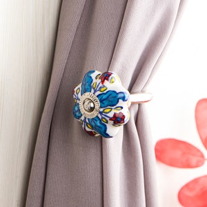 May include: A floral curtain tieback with a white, blue, and red design. The ceramic tieback is shaped like a flower and is attached to a gray curtain. The tieback has a silver metal ring.