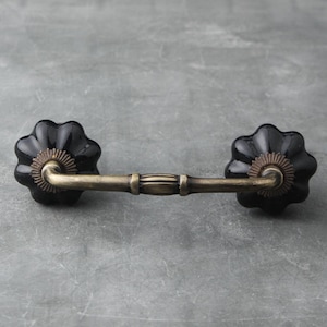 Pode incluir: Botões de armário de cerâmica preta com uma alça arqueada de metal dourado. Os botões têm uma borda festonada e um centro dourado.