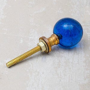 May include: A blue glass ball cabinet knob with brass hardware and a threaded rod. The knob has a round shape and is a vibrant shade of blue. The brass hardware includes a nut and washer.
