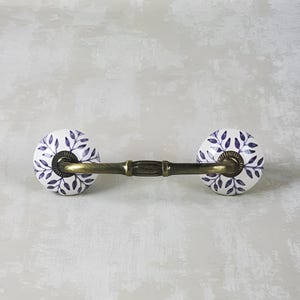 Pode incluir: Uma puxador de gaveta de cerâmica branca com um design floral roxo e uma barra de latão que conecta as duas maçanetas.