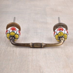 May include: A brass cabinet pull with ceramic knobs. The knobs are white with a yellow flower design and red accents.