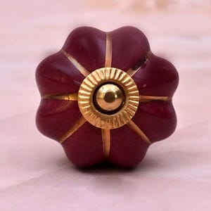Op de afbeelding: Een bloemvormige meubelknop met een diepe bordeauxrode, glanzende afwerking. De knop heeft zes afgeronde bloemblaadjes gescheiden door dunne, verdiepte goudkleurige lijnen. Een ronde, geribbelde goudkleurige metalen plaat zit in het midden.