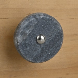 Może przedstawiać: Okrągła, szara kamienna gałka do szafki ze srebrną metalową śrubą. Gałka jest osadzona na jasnobrązowej drewnianej powierzchni, ukazując naturalne usłojenie drewna.