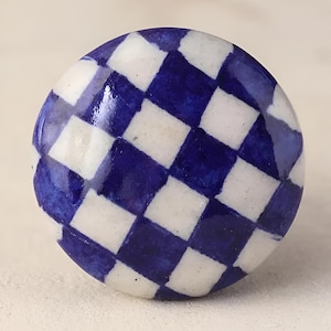 May include: A round, ceramic cabinet knob with a hand-painted checkerboard pattern in white and navy blue. The knob has a glossy finish and is designed for furniture or decorative use.
