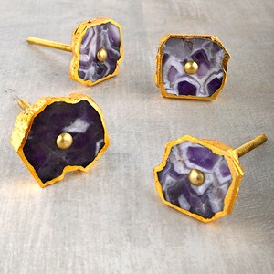 Może przedstawiać: Cztery ametystowe gałki do szafek z akcentami ze złoconego metalu. Każda gałka ma unikalny, nieregularny kształt i wzór w kolorze fioletowym i białym.