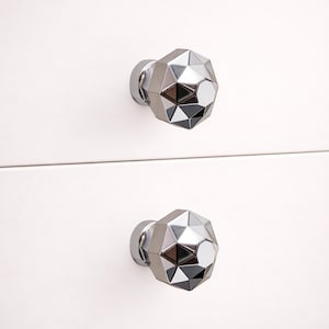 May include: Two silver geometric cabinet knobs with a faceted design are mounted on a white surface. The knobs have a modern, reflective appearance, adding a touch of elegance to any furniture piece. The knobs are made of metal.