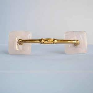 Op de afbeelding: Een gouden kastgreep met twee vierkante, doorschijnende witte knoppen.