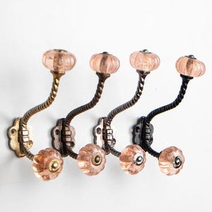 Op de afbeelding: Vier decoratieve haken met een gedraaid metalen ontwerp en roze glazen knoppen. De haken zijn gemonteerd op een witte muur.