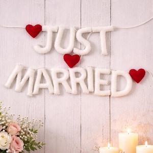 May include: A white felt banner with the words "JUST MARRIED" in fluffy letters, accented by red felt hearts. The banner hangs against a white wooden background, with flowers and candles in the foreground.