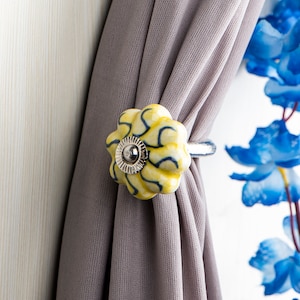 Pode incluir: Um amarrador de cortina de cerâmica amarelo e azul com um anel prateado. O amarrador tem a forma de uma flor com um padrão de espiral.
