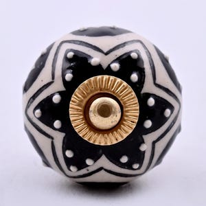 May include: A round ceramic cabinet knob featuring a black and white star-shaped pattern with small white dots. The centre of the knob has a circular gold-coloured metal hardware piece with a ridged edge and a smooth, flat centre point.
