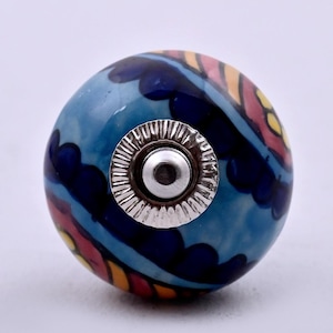 Puede incluir: Un pomo de cerámica redondo para muebles con acabado brillante y un patrón pintado a mano de formas curvas en azul, amarillo y rojo. El centro tiene un herraje de metal plateado con una placa base circular y estriada.