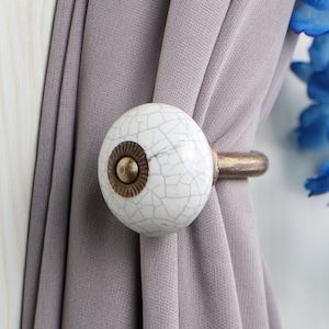 May include: White ceramic curtain tieback with a crackled finish and a gold metal ring. The tieback is attached to a gray curtain.