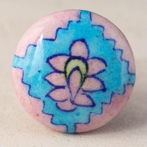 May include: A pink and blue ceramic knob with a floral design. The knob has a light pink background with a blue and white geometric pattern. In the centre of the knob is a pink flower with a yellow centre.