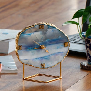 Puede incluir: Un reloj de ágata azul y blanco con detalles dorados y números romanos. El reloj está sobre un soporte de metal dorado.