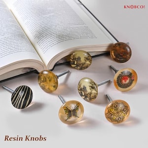 May include: A selection of resin knobs with diverse designs, such as a butterfly and a floral pattern. The knobs are presented on a white surface, with an open book in the background. The text "Resin Knobs" is visible at the bottom of the image.