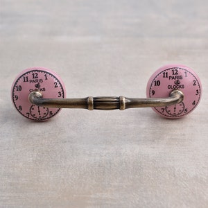 以下が含まれることがあります： 金色の金属棒で繋がれた、ピンクのセラミック製の引き出しノブ2個。それぞれのノブには、"Paris Clocks"という文字と黒いローマ数字の目盛りが付いた時計の文字盤があります。