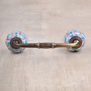 以下が含まれることがあります： 真鍮のバーと青と白のセラミックノブ2つが付いた装飾的なキャビネットプル。ノブはストライプ模様です。