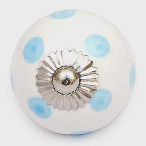 Könnte beinhalten: Ein runder weißer Keramik-Möbelknopf mit hellblauen Punkten. In der Mitte des Knopfes befindet sich ein silberfarbenes Metallteil mit einem gezackten, sonnenstrahlenartigen Rand und einer kleinen zentralen Wölbung als dekorativer Abschluss.