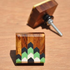 May include: Two wooden cabinet knobs with a geometric design. The top knob is brown with a yellow top and a silver screw. The bottom knob is brown with a green, white and black geometric design.