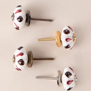 Könnte beinhalten: Vier weiße Keramik-Schrankknöpfe mit rotem und braunem Blumendesign. Jeder Knopf hat eine Schraube in einer anderen Farbe.