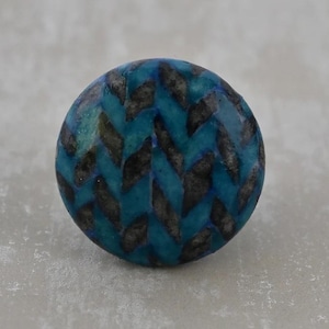 May include: A round, decorative knob with a teal and black chevron pattern. The knob is a solid color with a glossy finish. The chevron pattern is made up of black, elongated shapes that are arranged in a zig-zag pattern.