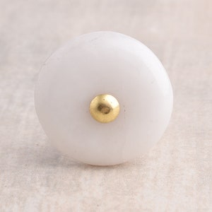 Op de afbeelding: Witte keramische knop met een gouden midden. De knop is rond en heeft een glad oppervlak.