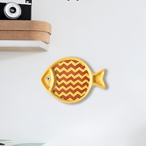 May include: A yellow ceramic fish-shaped dish with a brown and yellow chevron pattern. The dish has a small, white eye.