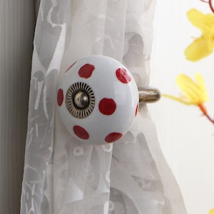 Könnte beinhalten: Weißer Vorhang-Halter aus Keramik mit roten Punkten und einem silbernen Metallhaken.