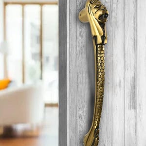 Könnte beinhalten: Ein goldener Türgriff mit einem Löwenkopf und einem strukturierten, gebogenen Körper. Der Griff ist an einer weißen Holztür montiert.