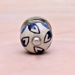 May include: A round ceramic cabinet knob with a cream-colored base featuring a hand-painted pattern of dark blue teardrop shapes outlined in black. The knob has a central silver-toned metal screw head and a silver-toned metal base plate.